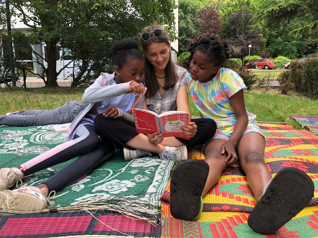 Une adulte lit un livre à deux fillettes, installées par terre dans un parc.