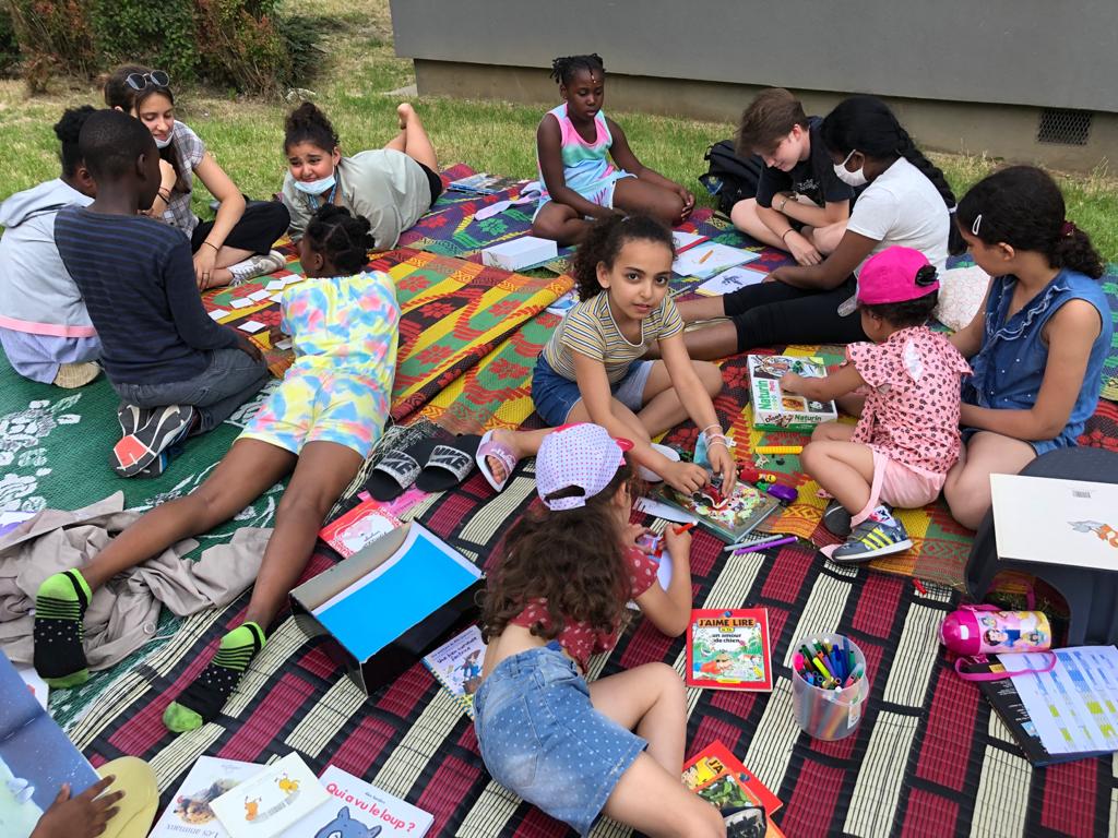 Des enfants lisent ou discutent, installés sur un tapis coloré, dans un parc
