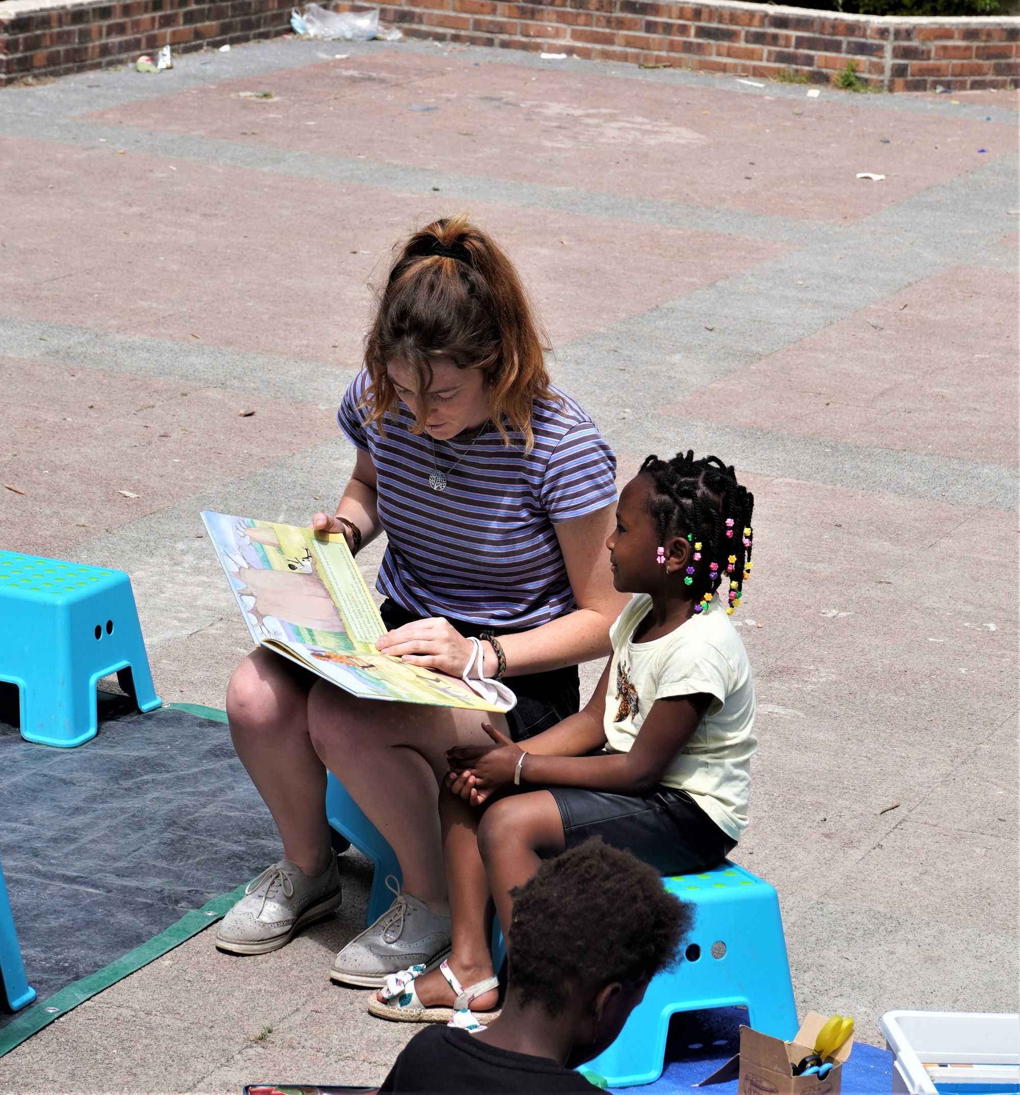 Une adulte lit un livre à une fillette. Extérieur.